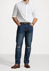 Man draagt een wit overhemd met knopen, blauwe versleten spijkerbroek, bruine leren riem, bruine loafers met kwastjes, met handen in de zakken tegen een effen achtergrond.