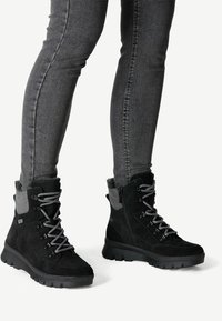 Botas negras hasta el tobillo hechas de gamuza y material sintético, con acentos grises y suelas gruesas y texturizadas. Son visibles los cordones y cremalleras.