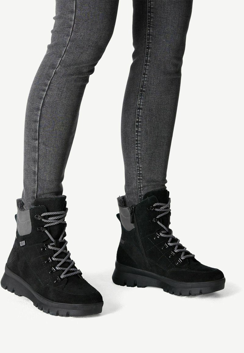Botas negras hasta el tobillo hechas de gamuza y material sintético, con acentos grises y suelas gruesas y texturizadas. Son visibles los cordones y cremalleras.