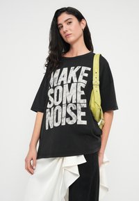 Μαύρο φαρδύ μπλουζάκι με λευκό κείμενο "MAKE SOME NOISE". Διαθέτει υφή υφάσματος. Συνδυάζεται με σκούρο παντελόνι και πράσινη τσάντα με υφή ιμάντα.