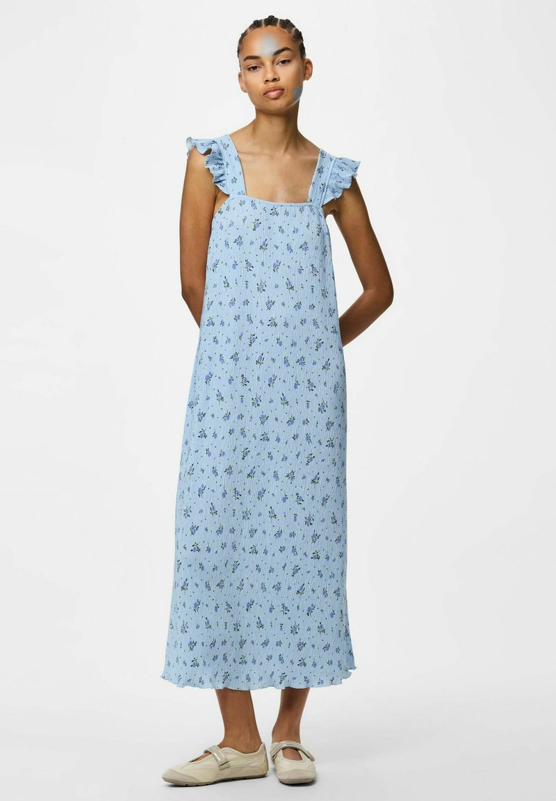Femme portant une robe midi fleurie bleu clair sans manches avec des bretelles à volants et des chaussures plates de couleur crème, debout les mains dans le dos.