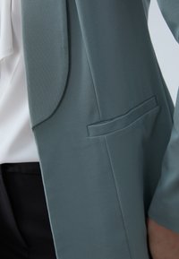 Veste bleu clair texturée avec une finition douce, présentant une coupe slim, une poche unique et un chemisier blanc lisse en dessous.