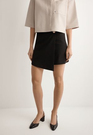 Kobieta w beżowej krótkiej koszuli, czarnej asymetrycznej mini spódniczce z złotym guzikiem oraz czarnych butach na blokowym obcasie typu slingback.