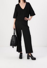 Femme portant un pantalon noir à jambes larges, un pull noir à col en V, des mocassins noirs à talons larges, tenant un sac fourre-tout en cuir noir, debout.