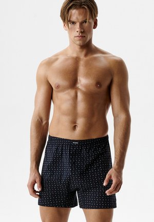  umięśniony mężczyzna z jasnobrązowymi włosami ubrany w czarne bokserki z wzorem, stojący na tle jednolitej białej ściany.