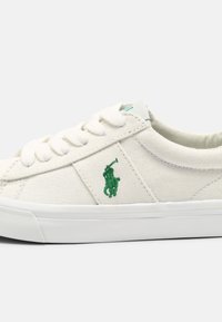 Zapatilla de lona blanca con cordones blancos y logo de jugador de polo bordado en verde en el lado, con suela de goma blanca.