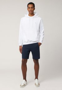 Sweatshirt à capuche en coton blanc avec poche avant, associé à un short bleu marine. Des baskets décontractées avec des accents beige et blancs complètent le look.