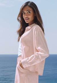Light pink button-up shirt made from soft fabric, featuring a relaxed fit and long sleeves. Worn over a white top and paired with matching pants.