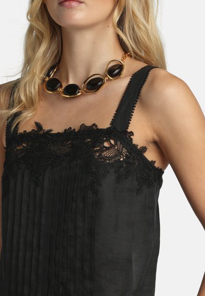 Femme blonde portant un haut sans manches noir orné de dentelle et un collier épais en or avec de grandes pierres ovales noires.