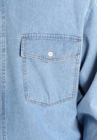 Jasnoniebieska koszula dżinsowa z kieszenią z zapięciem na guziki i widocznymi szwami na klatce piersiowej, prezentująca teksturę materiału i szwy.