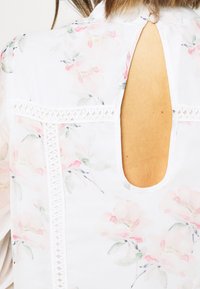 White floral blouse featuring soft pink and green watercolor patterns, a keyhole back design, and intricate lace detailing at the seam.