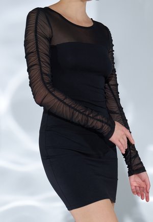 Femme portant une robe noire ajustée avec de longues manches transparentes froncées, debout devant un fond clair avec des motifs d'ombres douces.