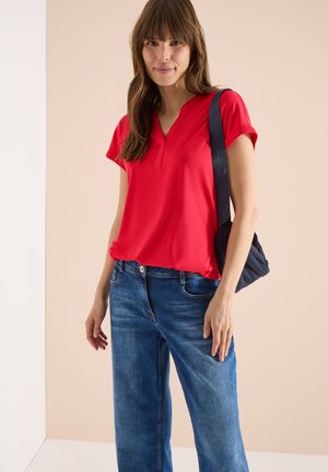 Femme portant un t-shirt rouge à manches courtes, un jean bleu, et un sac à main noir sur une épaule, sur un fond beige.