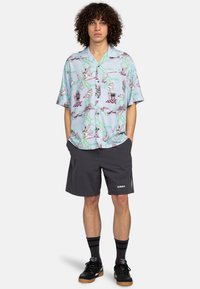 Kurzarmhemd in Hellblau mit buntem Grafikdruck, kombiniert mit dunkelgrauen Shorts. Schwarze Sneakers und gestreifte Socken runden den Look ab.
