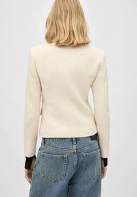 Chaqueta ajustada color crema con un acabado texturizado, que presenta mangas largas y un diseño estructurado, combinada con jeans azules de cintura alta.