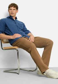 Blaues Hemd mit Knöpfen, einem strukturierten Muster und langen Ärmeln, kombiniert mit braunen Chinos und weißen Sneakern, sitzend auf einem Chromstuhl.