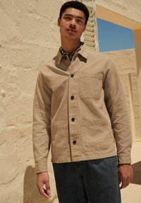 Beige Utility-Jacke aus strukturiertem Stoff, mit Knopfleiste, aufgesetzter Tasche und zwei unteren Taschen, kombiniert mit dunkelblauen Jeans.