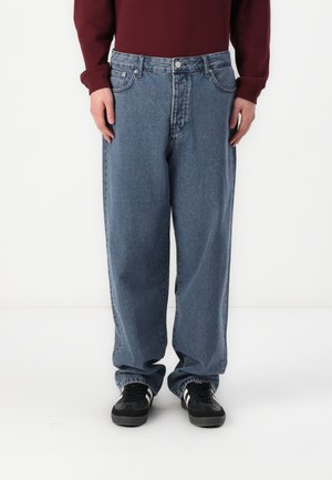 Light blue, loose-fitting denim jeans with a button and zipper fly, five pockets, and contrast stitching, worn with black sneakers.