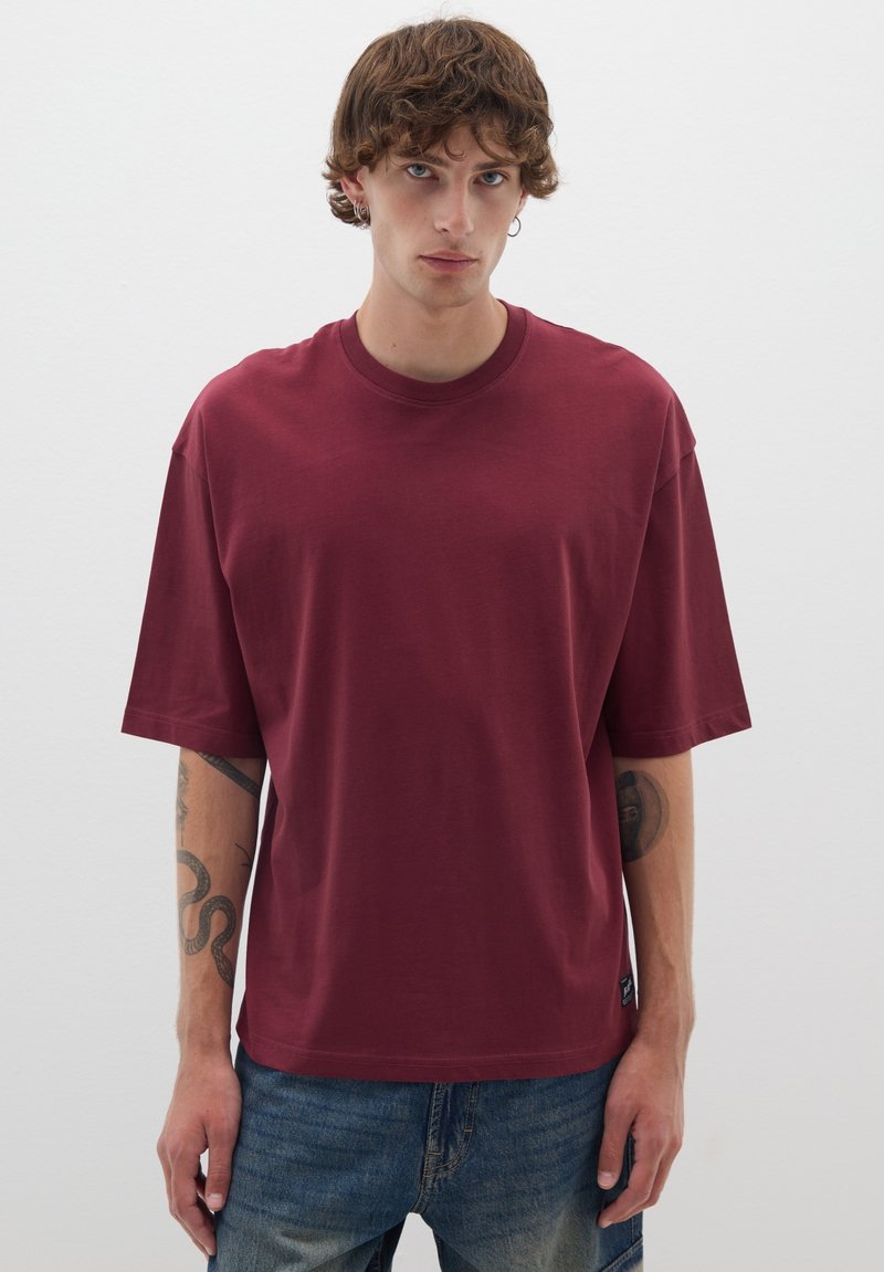 T-shirt oversize borgogna realizzata in cotone. Maniche corte, scollo rotondo e una texture morbida. Abbinata a jeans blu chiaro.