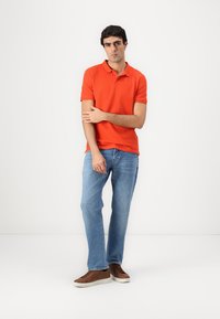 Camisa de polo naranja con cuello, combinada con jeans azul claro y zapatos marrones. La tela parece suave, con un diseño ajustado.