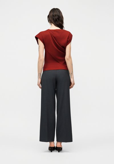 Γυναίκα με σκούρα κυματιστά μαλλιά, που φοράει κόκκινο κοντομάνικο τοπ και σκουρόχρωμο παντελόνι με φαρδιά πόδια, στέκεται γυρισμένη προς τα πίσω πάνω σε λευκό φόντο.