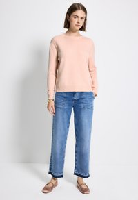 Jeune femme portant un pull rose clair, un jean bleu bicolore avec de grandes poches avant, et des ballerines rose, se tenant devant un fond blanc.