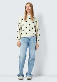 Γυναίκα που στέκεται φορώντας κρεμ πουλόβερ με μαύρο σχέδιο καρδιάς, ανοιχτό μπλε τζιν και λευκά αθλητικά παπούτσια, μπροστά σε απλό γκρίζο φόντο.