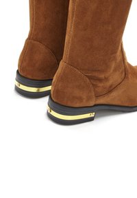 Braune Wildleder-Stiefel bis zum Knie mit runder Zehe und schwarzem Gummisohle. Verfügt über einen goldenen Akzentstreifen und dekorative Beschläge in der Nähe der Ferse.