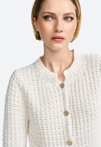Cardigan blanc en tricot avec un motif texturé, encolure ronde et trois boutons décoratifs en ton doré. Tissu doux et léger.