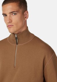 Brun höghalsspets med kvartdragkedja, med ribbad textur och subtil kontrastsöm längs axlarna och halslinningen.