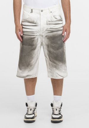 Hombre con pantalones cortos hasta la rodilla en degradado blanco y gris, calcetas blancas y zapatillas negras y blancas, de pie con las manos relajadas a los lados.