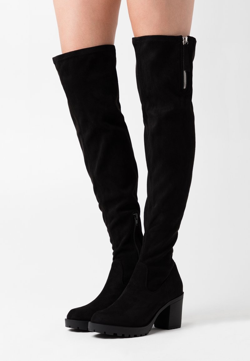 Botas de gamuza negra por encima de la rodilla con un tacón grueso, que cuentan con cremalleras laterales y una textura suave, diseñadas para ajustarse perfectamente a la pierna.