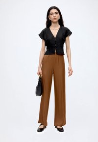 Femme aux longs cheveux foncés portant un haut noir à manches courtes boutonné, un pantalon large marron, des chaussures noires, et tenant un sac à main noir.