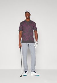 Man wearing a gray polo with purple golf club patterns, light gray pants, white golf glove, cap, and holding a golf club against a plain background.
