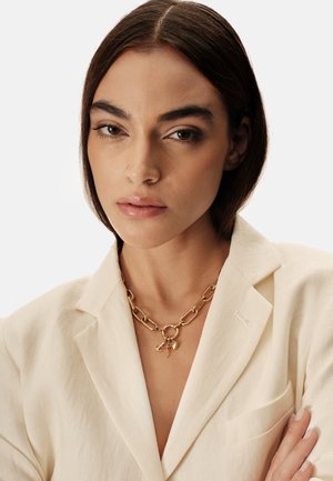 Gouden ketting met grote schakels en een uniek bedelhangertje, gedragen boven een beige blazer met een subtiele structuur.