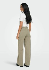 Femme aux longs cheveux noirs portant une chemise blanche à manches courtes, un pantalon beige à jambes larges et des chaussures noires, debout sur un arrière-plan neutre.