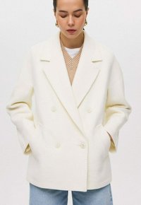 Dubbelzijdige witte jas van een dikke stof met ingesneden revers, twee knopen, zijzakken en een ontspannen pasvorm. Gelaagd over een beige trui.