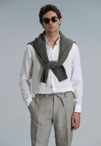 Chemise en lin légère de couleur blanche, associée à un pantalon rayé beige. Un pull en maille vert foncé drape sur les épaules. Lunettes de soleil rondes portées.