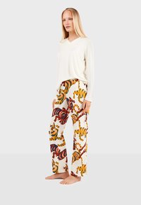 Lichtbeige lange mouwen v-hals top gecombineerd met een broek met wijde pijpen, versierd met een kleurrijk tijgerpatroon op een off-white achtergrond.