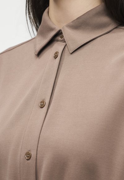 Κοντινό πλάνο ενός ατόμου που φοράει μια πουκάμισα ταυ πέιτζ καφέ με κουμπιά και μυτερό γιακά, με ορατά καφέ κουμπιά.