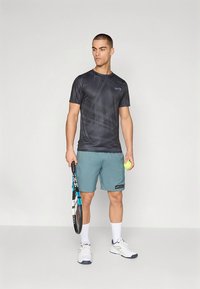 Svart mönstrad sportt-shirt och tealfärgade shorts. Håller i en tennisklubb och boll, vita sneakers med marinblå detaljer, står på en neutral bakgrund.