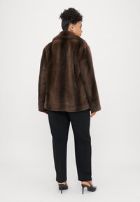 Brązowy płaszcz z sztucznego futra z kołnierzem, o luźnym kroju i długich rękawach, zestawiony z czarnymi spodniami o dopasowanym fasonie oraz czarnymi butami na obcasie.