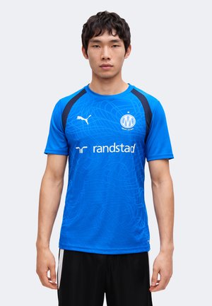 Uomo che indossa una maglia sportiva Puma blu con accenti neri, logo del club di calcio di Marsiglia e nome dello sponsor Randstad, in piedi davanti a uno sfondo semplice.