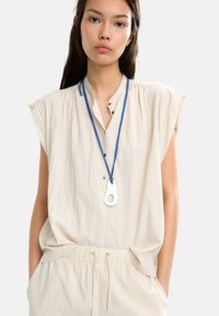Beżowa, bezrękawnikowa bluzka z zapięciem na guziki. Posiada niebieski sznur i srebrny wisiorek z owalnym wycięciem. Miękki, teksturowany materiał.