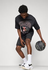 Črna majica s kratkimi rokavi z grafiko "Chicago Bulls", črne mrežaste kratke hlače, bele nogavice in črne ter bele športne čevlje. Drži košarko.