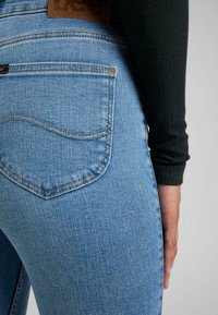 Jeans de denim azul con un primer plano del bolsillo trasero. La tela tiene una textura suave, con costuras en contraste y una etiqueta de marca de cuero marrón.