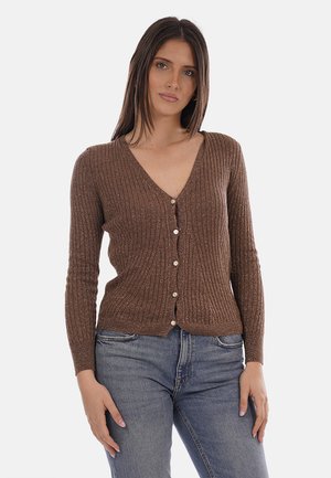 Donna con capelli castani lisci che indossa un cardigan marrone a coste con bottoni e jeans blu, in piedi con le mani rilassate ai lati.