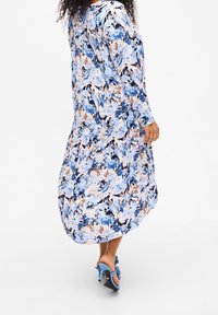 Květinové šaty s dlouhým rukávem, které mají modré, béžové a černé vzory; lehká látka volně splývá; v kombinaci s modrými sandály na vysokém podpatku.