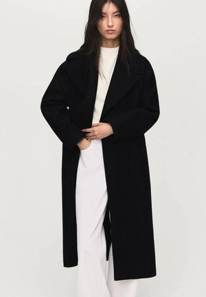 Cappotto di lana nera con un ampio colletto, maniche lunghe e silhouette dritta; il modello lo indossa sopra un top chiaro e pantaloni bianchi.