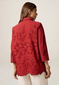 Rote bestickte Bluse mit aufwendigen Ausschnittdesigns, dreiviertellangen Ärmeln und abgerundetem Saum. Strukturierter Baumwollstoff.